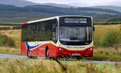 A red and cream single-decker bus with ‘West Coast Motors’ branding is travelling on a rural road surrounded by green fields and rolling hills. The bus has large windows, a front destination display, and a registration plate reading SJ23 HRP. The background shows distant hills under a cloudy sky.