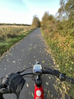 The picture has been taken over the handlebars of a red and black bike. There is a cycle path in front, with a hedge on either side. The sky ahead is blue.