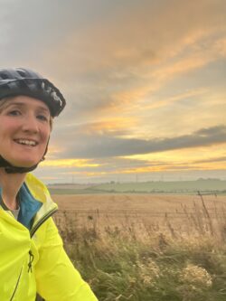 A female wearing a cycle helmet and a yellow high vis jacket with some fields in the background and a pretty sunset sky.