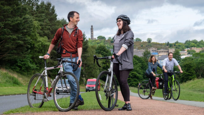 Two people in the foreground, each standing next to a bike. Behind them and to the right are two people cycling on a cycle path. To the left of the people is a road, with trees next to it. Edinburgh can be seen in the background.
