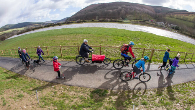 a group of children and adults are cycling and scooting along a new path. There is grass is the foreground, followed by path, more grass, a loch and then a hill.