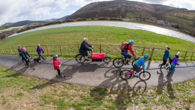 a group of children and adults are cycling and scooting along a new path. There is grass is the foreground, followed by path, more grass, a loch and then a hill.