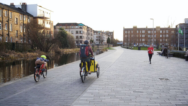 An adult and child are cycling away from the camera on a wide, paved cycleway. On their left is a river or canal and on the left of this are flats. The adult is riding a cargo bike with a large yellow container on the front. the child is riding a red bike.