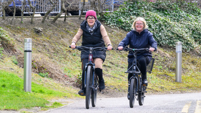 Two people cycling along a cycle path. There is grass to their left and in the background a car park.