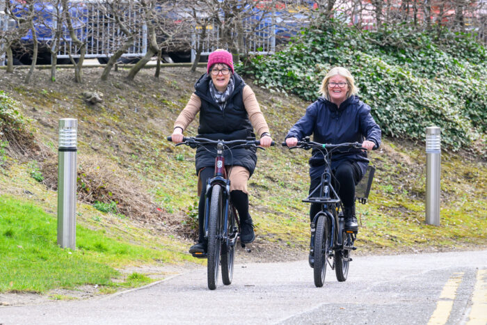 Two people cycling along a cycle path. There is grass to their left and in the background a car park.