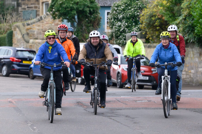 A group of people cycling along a road. There are parked cards behind them on the right.