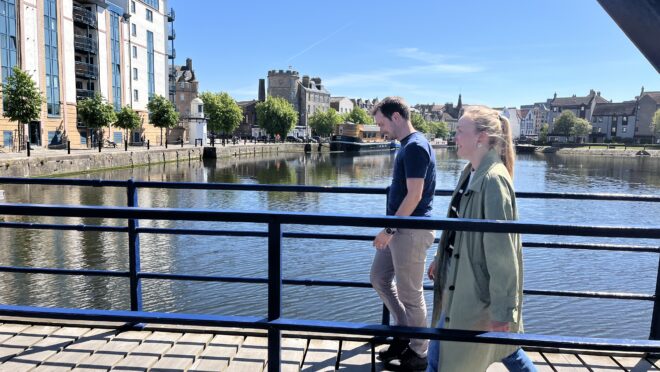 Two people walking from right to left. There is water behind them and a clear blue sky. To the left are blocks of flats.