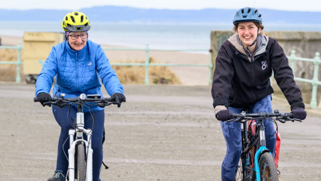 Two people cycling towards the camera. They are on a road or pavement and there is a beach and the sea visible in the background.