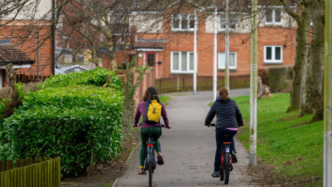 Two people cycling away from the camera on a cycle path. There are hedges on either side and houses in the distance.