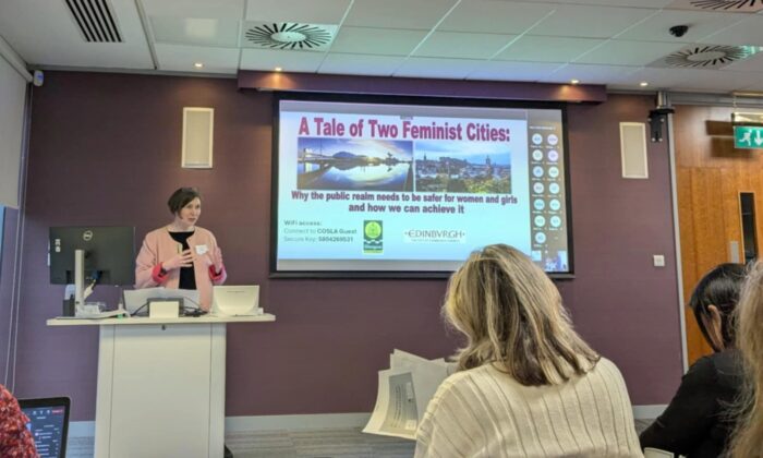 A woman is presenting at the front of a room. there is a screen on the wall which says 'a tale of two feminist cities'.