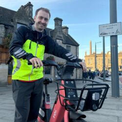 A man wearing a yellow high vis jacket standing with a bike on a pavement with a building in the background.