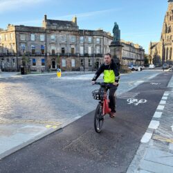 A man wearing a yellow high vis jacket cycling along a designated cycle land with a road and buldings to the left.