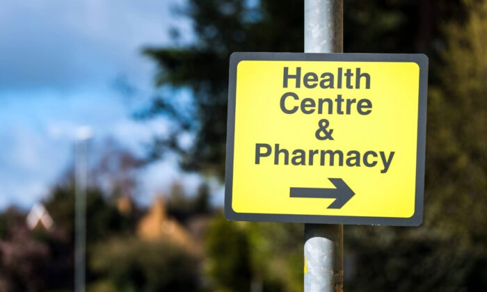 Yellow directional sign reading 'Health Centre & Pharmacy' with a right-pointing arrow, mounted on a metal pole against a blurred outdoor backdrop of trees and buildings.