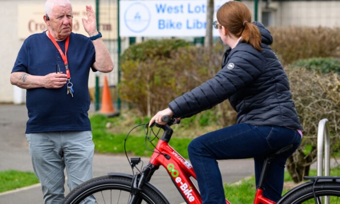 An older person with short white hair, dressed in a navy blue shirt and light grey trousers, stands gesturing with one hand and holding keys in the other. A younger person with long hair tied back, wearing glasses, a black jacket, and jeans, sits on a red bicycle labelled 'GO e-Bike'. They are outside near a building with a sign reading 'West Lothian Bike Library', with greenery and part of the building visible in the background.