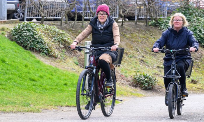 Two individuals ride upright bicycles on a paved path outdoors. The person on the left wears a red hat, glasses, a black vest over a beige long-sleeve shirt, and black trousers. The person on the right wears glasses and a dark blue jacket with black trousers. The background features green grass, bushes, and parked cars behind a fence.