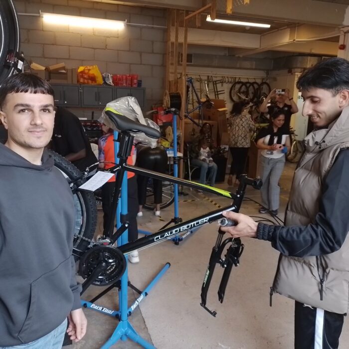 A person works on a bicycle held in a repair stand inside a busy workshop, while another person stands beside them. Several people and bikes are visible in the background.