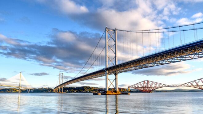 A large suspension bridge spans calm water under a bright sky with scattered clouds. To the left, a modern cable‑stayed bridge rises in pale tones, while a red cantilever bridge appears in the distance on the right. The sunlight highlights the metal structures and reflects softly on the water.