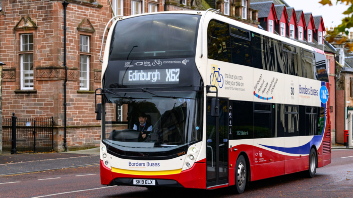 A double decker Borders Buses service in red, white and blue livery travelling on a city street. The front destination display shows Edinburgh X62, and the bus passes stone buildings and autumn trees.