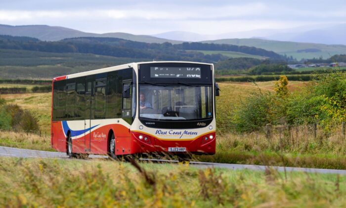 A single decker bus travelling along a rural road, surrounded by grassy fields and rolling hills, with a cloudy sky and distant countryside in the background.