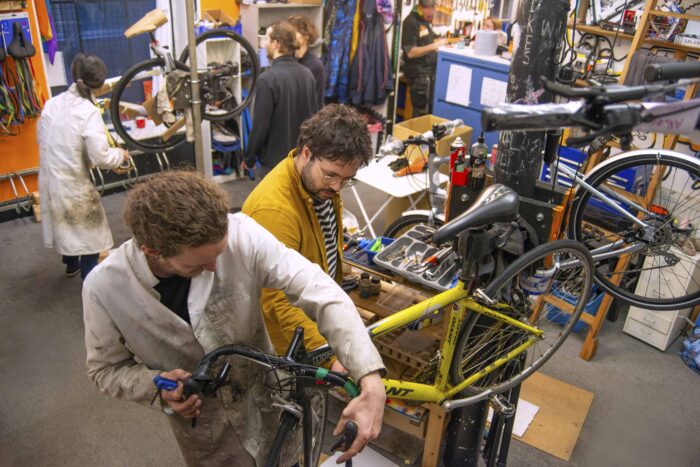 People repairing bicycles in a busy workshop, with bikes mounted on stands and tools laid out on workbenches.
