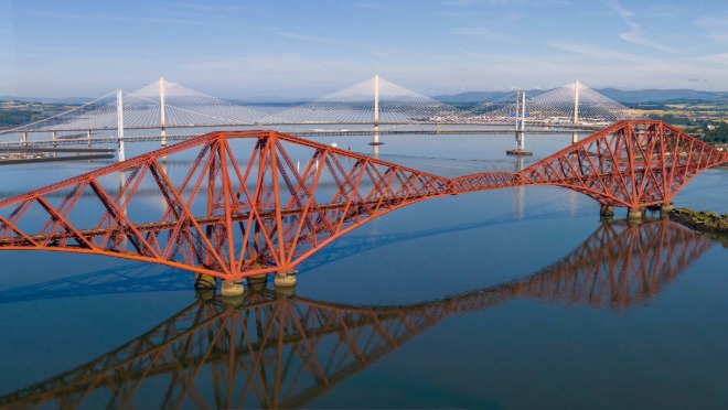 Aerial view of the red Forth Bridge with the Forth Road Bridge and Queensferry Crossing behind it, spanning the Firth of Forth on a clear day.