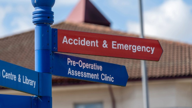 Directional sign outside a hospital showing arrows for Accident and Emergency and Pre‑Operative Assessment Clinic, with a building in the background.