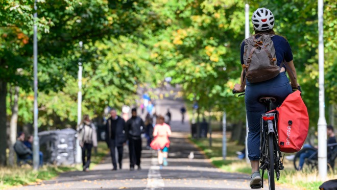 Person cycling along a tree‑lined shared path, carrying a red pannier bag, with pedestrians walking ahead in the distance.