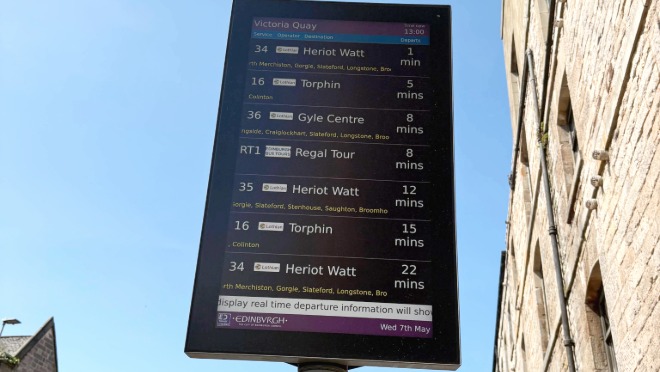 Digital bus stop display showing live departure times for routes from Victoria Quay, including services to Heriot Watt, Torphin and Gyle Centre, mounted on a pole beside a stone building.