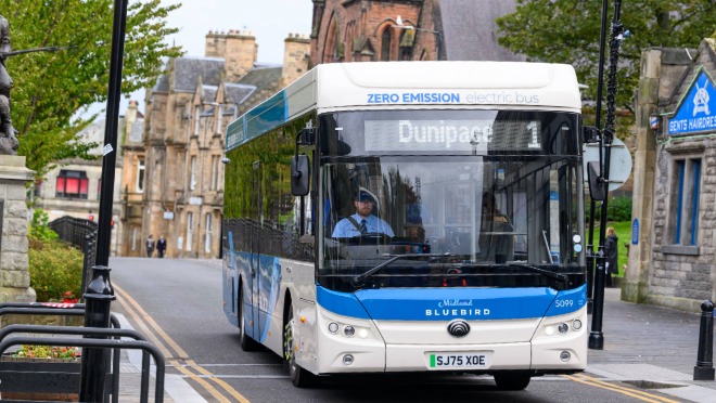 Electric bus on route 1 to Dunipace driving through a Scottish town, with stone buildings lining the street.