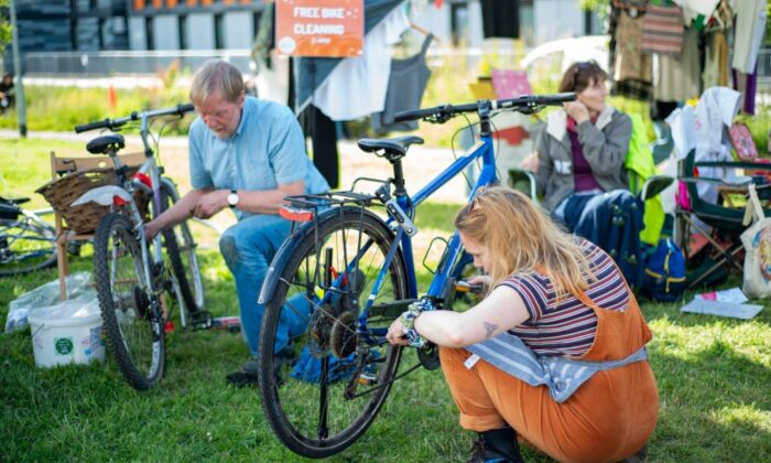 A person crouches on grass repairing a bicycle wheel at an outdoor bike repair event, with other bikes and people nearby.