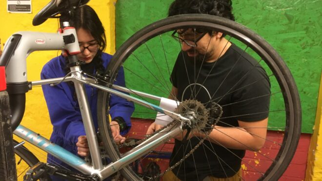 Two people working together to repair a bicycle mounted on a stand, focusing on the rear wheel and chain.