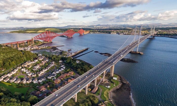 Aerial view of a road bridge crossing a wide river, with a red rail bridge nearby and housing along the shoreline.