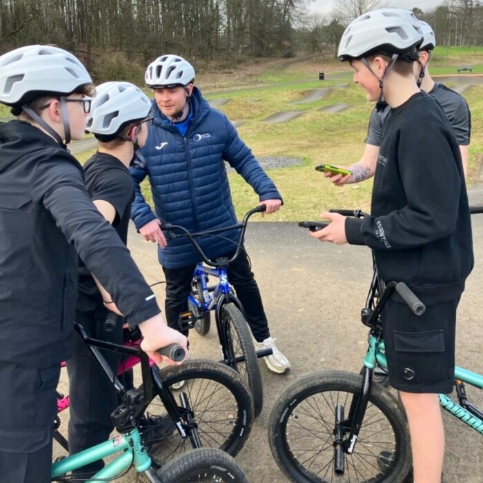 Group of young people wearing helmets standing with BMX bikes while an adult gives guidance at an outdoor track