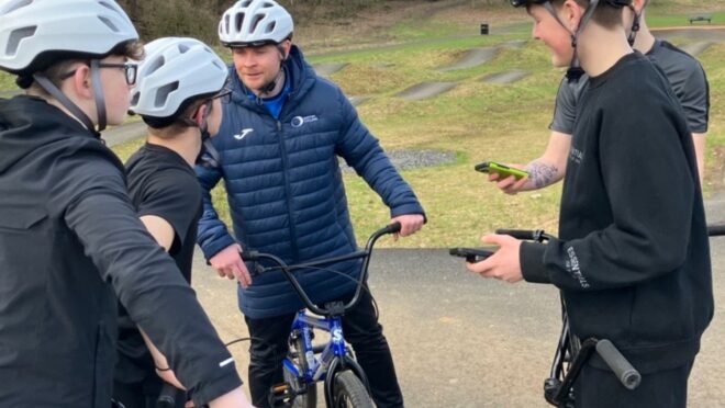 Group of young people wearing helmets standing with BMX bikes while an adult gives guidance at an outdoor track