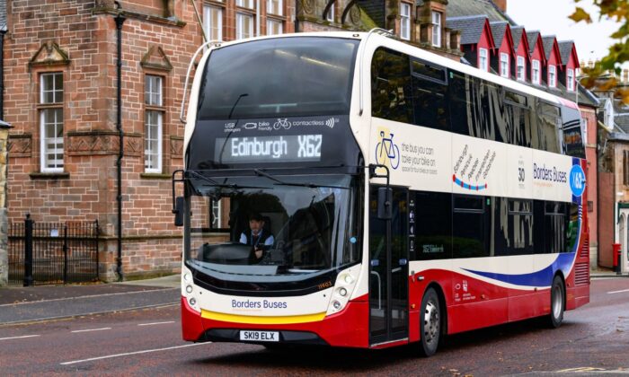 Red and white double-decker bus labelled “Borders Buses” driving along a street, with a digital display showing “Edinburgh X62,” passing a stone building and colourful houses in the background.