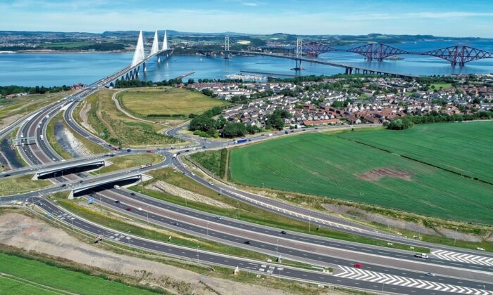 Aerial view of the Forth crossings in Scotland, showing the Queensferry Crossing, the Forth Road Bridge and the Forth Bridge spanning the Firth of Forth. Multi-lane roads and interchanges lead to the bridges, with surrounding farmland, housing and coastline visible on a clear day.