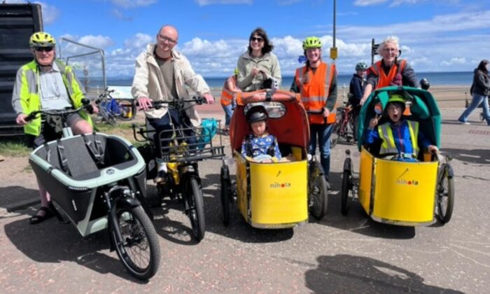 Participants on a variety of cycles and adapted bikes gather outdoors at a cycling event. In the centre, a person rides a yellow-fronted cargo bike carrying a child, while other riders on tricycles and bicycles wait nearby. A waterfront and blue sky are visible in the background.