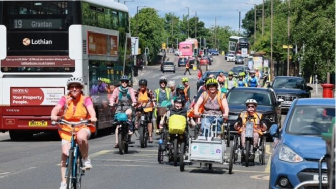 A group of cyclists ride along a busy urban road, travelling alongside cars, buses and other traffic. Several riders are on cargo bikes and adapted cycles, with a red double-decker bus visible in the background and trees lining the street.