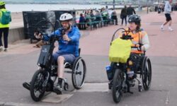 Two people ride adapted cycles along a waterfront path. In the foreground, one rider uses a handcycle while another rides a tricycle carrying a bright yellow bag. Additional cyclists and railings line the path in the background beside the water.
