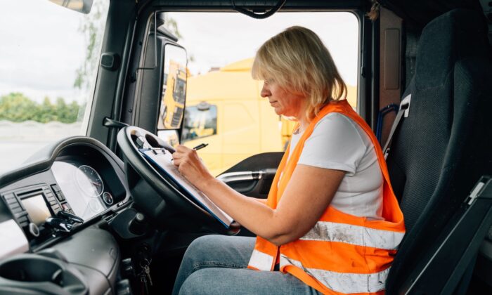 Person wearing a high-visibility orange vest sits in the driver’s seat of a lorry cab, writing on a clipboard. The vehicle dashboard and steering wheel are visible in the foreground, with another lorry seen through the window in the background.