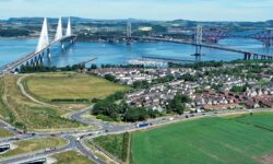 Aerial view of the Queensferry Crossing and Forth Road Bridge crossing the Firth of Forth, with houses, roads and farmland in the foreground.