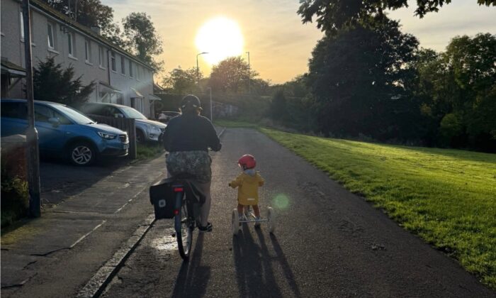 An adult cycles alongside a young child on a tricycle, both travelling along a quiet path beside homes and a grassy park as the low morning or evening sun casts long shadows ahead.