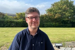 Person wearing glasses and a dark blue shirt standing outdoors in a grassy area. Trees and shrubs are visible in the background under a bright sky.