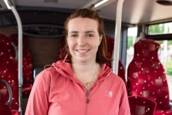 Person in a pink hooded jacket standing inside a bus, facing the camera. Red patterned bus seats and handrails are visible in the background, with daylight coming through the windows.