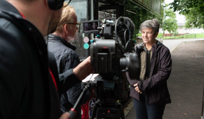 A person being filmed beside a bus, with a camera and two members of the filming crew visible in the foreground.